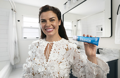 Smiling woman in a bright bathroom holds a tube of Enamūr toothpaste