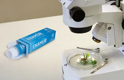 A tube of Enamür cream in its box lies next to a microscope and a petri dish containing green plant samples