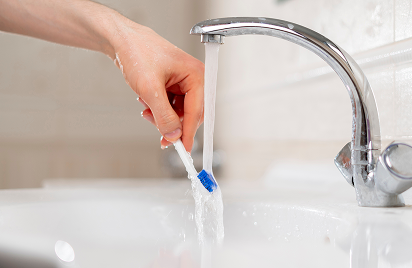 A hand holds a blue toothbrush under a running faucet in a white sink