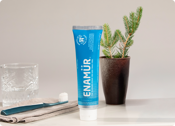 Tube of Enamür Peppermint toothpaste stands on a surface alongside a glass, toothbrush, folded towel, and potted plant
